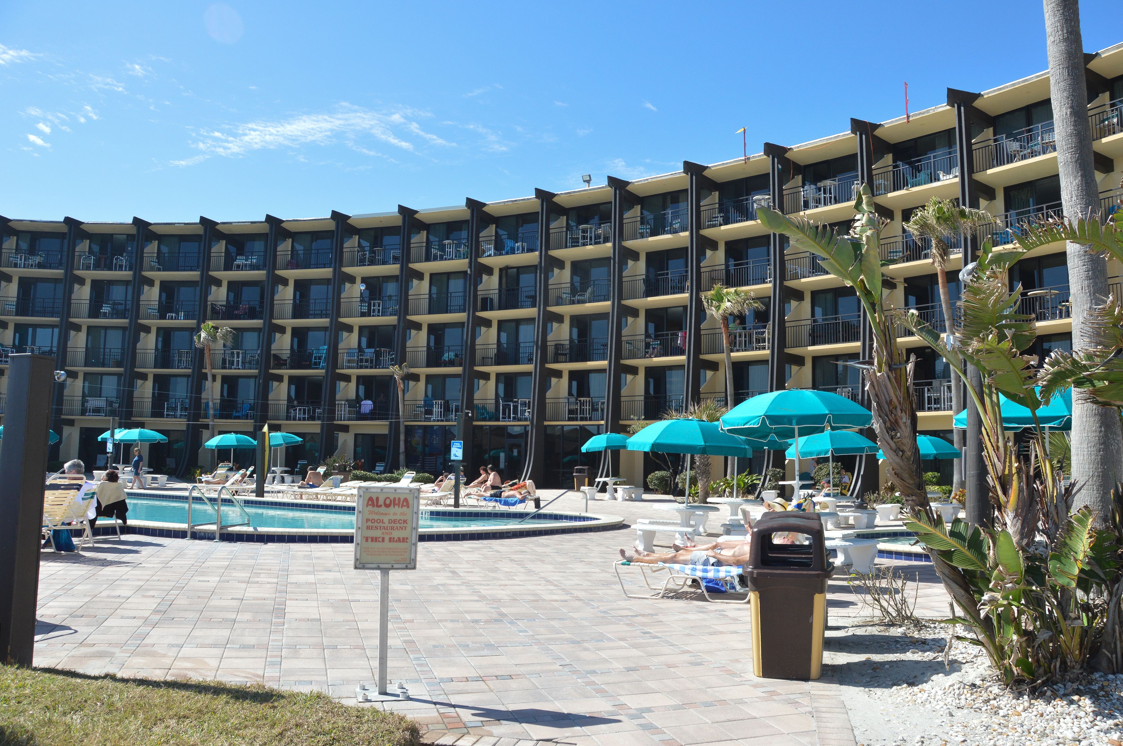Un hotel con zona de piscina, varios balcones y mobiliario exterior bajo sombrillas azules.