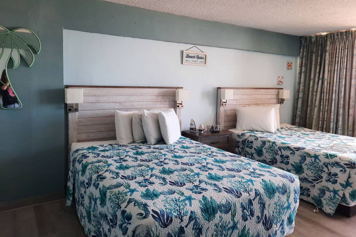 A hotel room with two beds, each with floral-patterned bedding, a small bedside table with a lamp, and a wall-mounted sign that reads "Beach Rules."