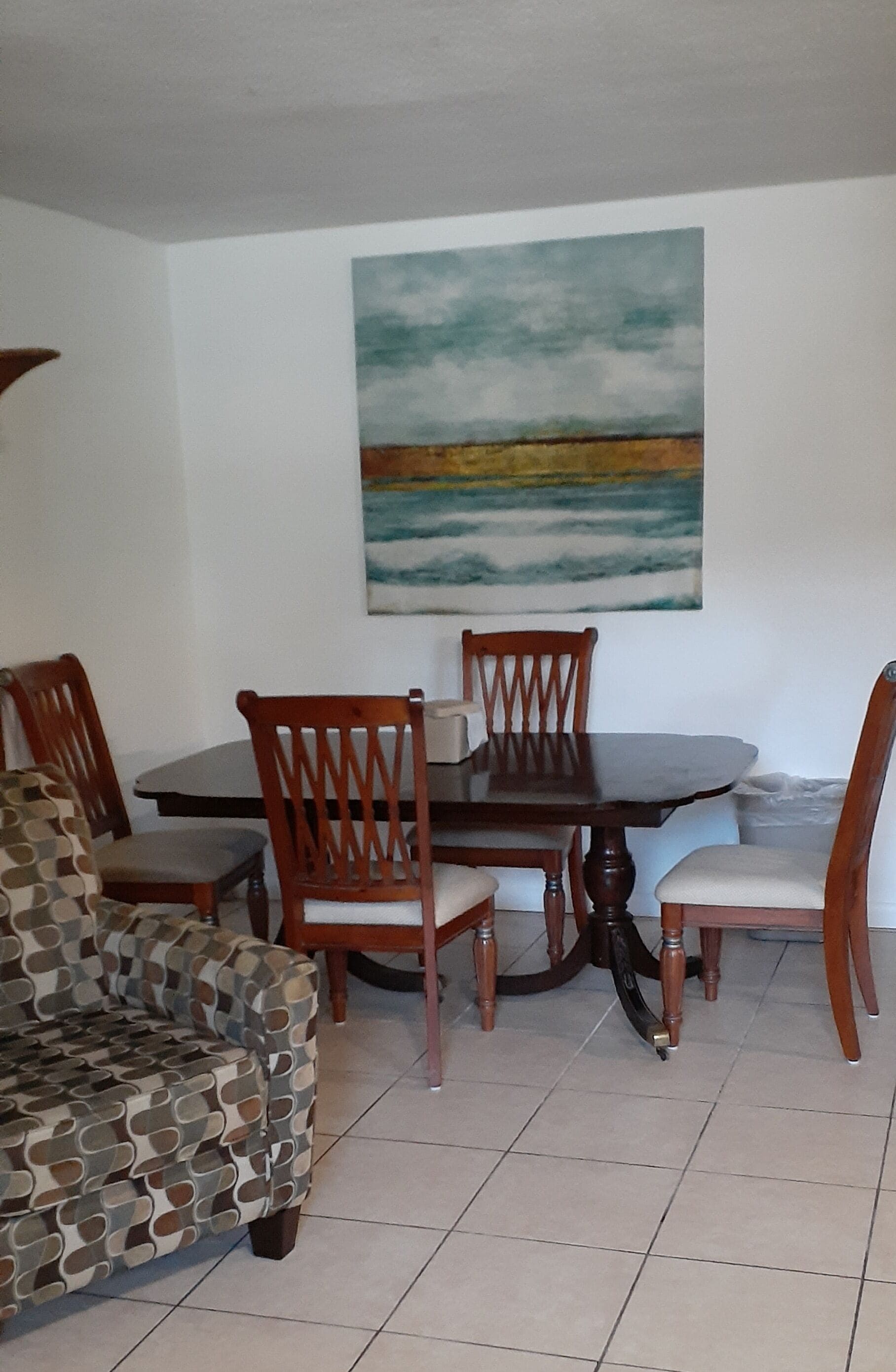 Un coin repas avec une table ronde et quatre chaises en bois, un canapé à motifs et un tableau représentant un paysage accroché au mur.