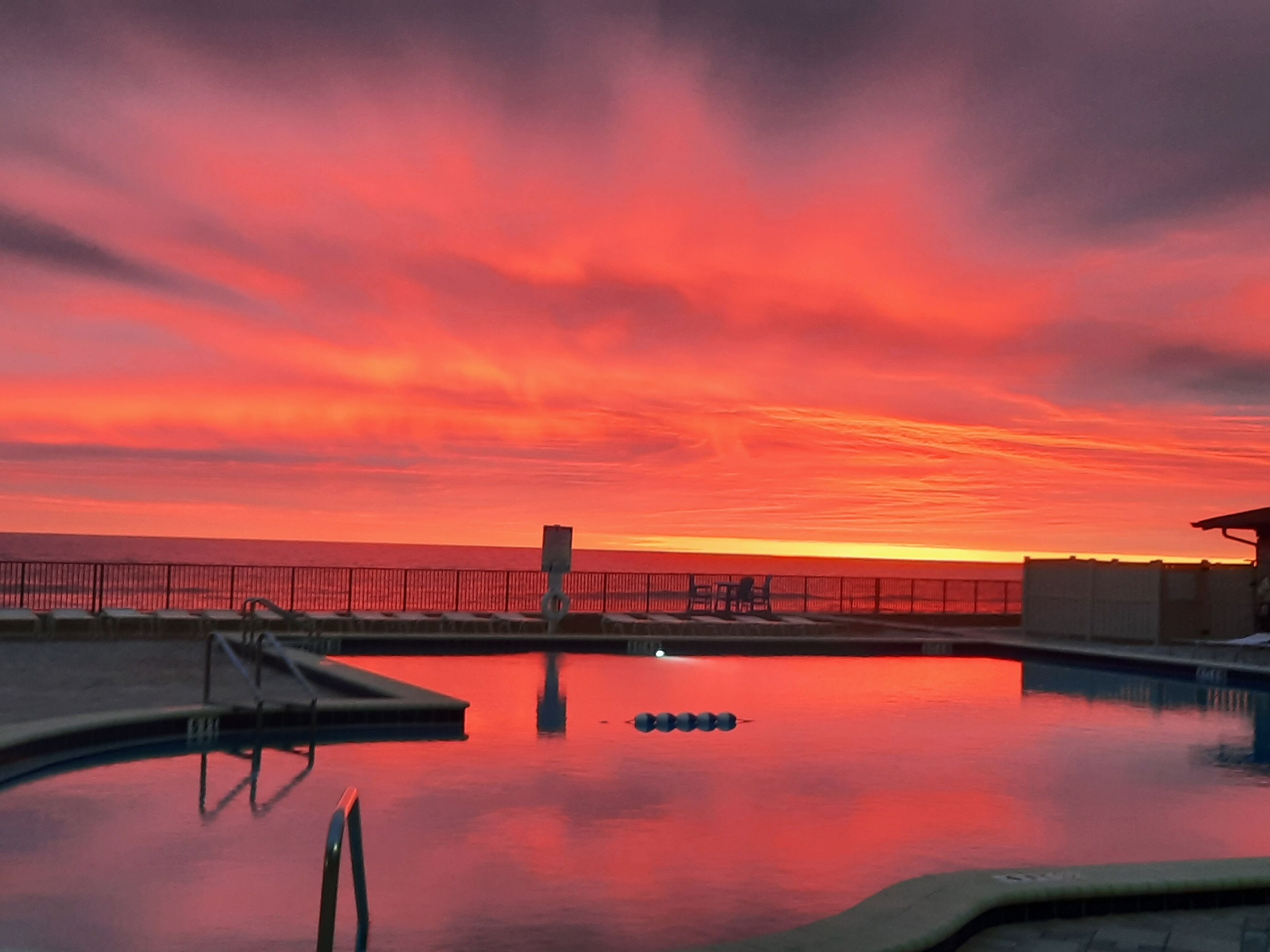 Un espace piscine paisible avec vue sur le coucher de soleil, une clôture et un bâtiment en arrière-plan.