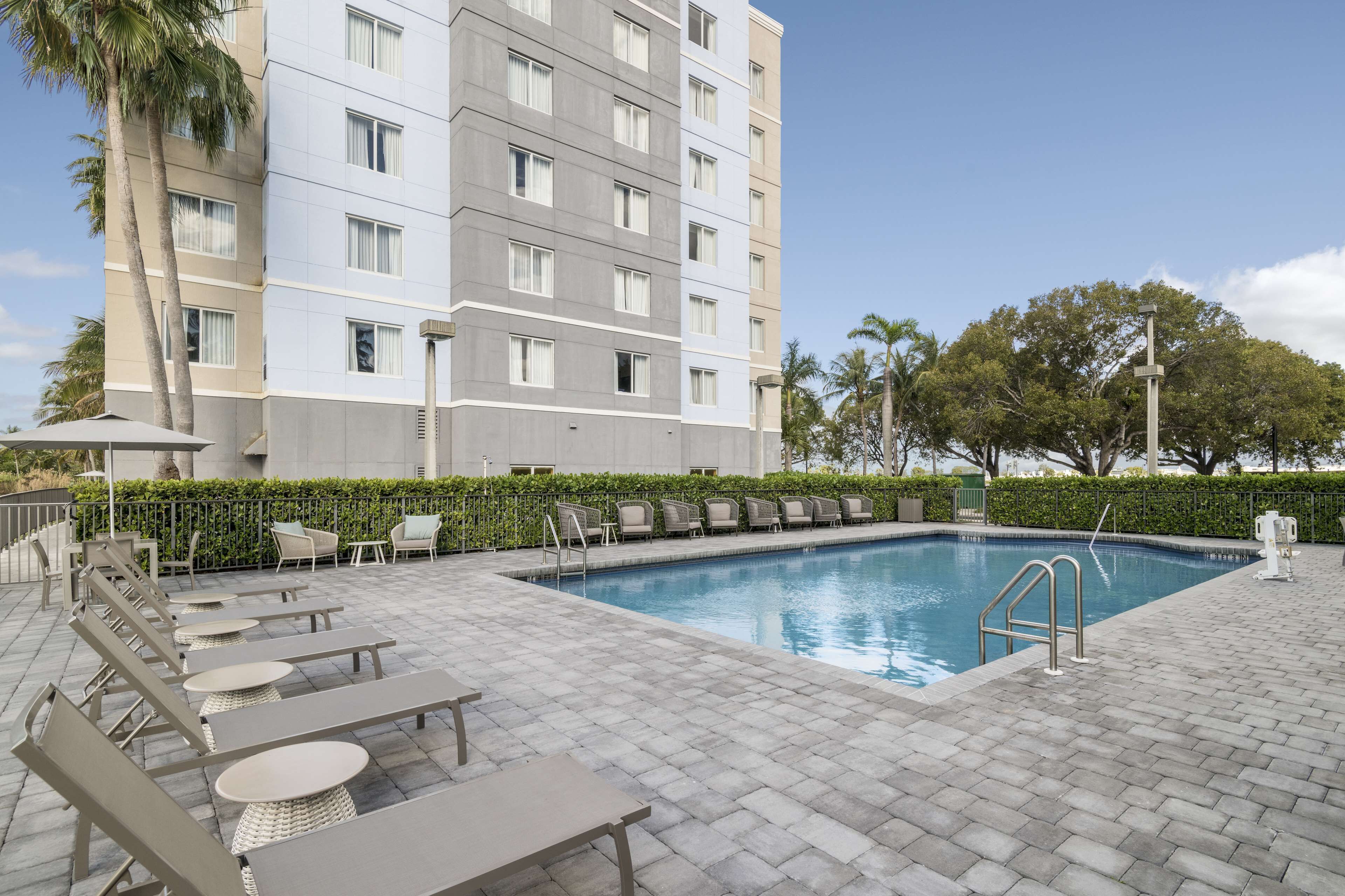 Zona de piscina de hotel con sillas de descanso y mesas, una piscina y un edificio con varios pisos y ventanas.
