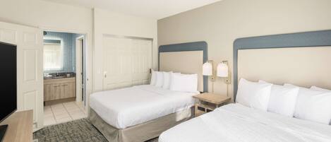 A hotel room with two beds, a television, a desk, and a kitchenette.