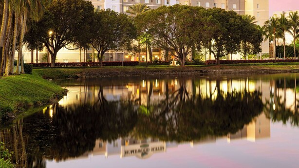 A hotel named Homewood Suites by Hilton with a reflection in a calm water body, surrounded by trees and well-maintained landscaping.