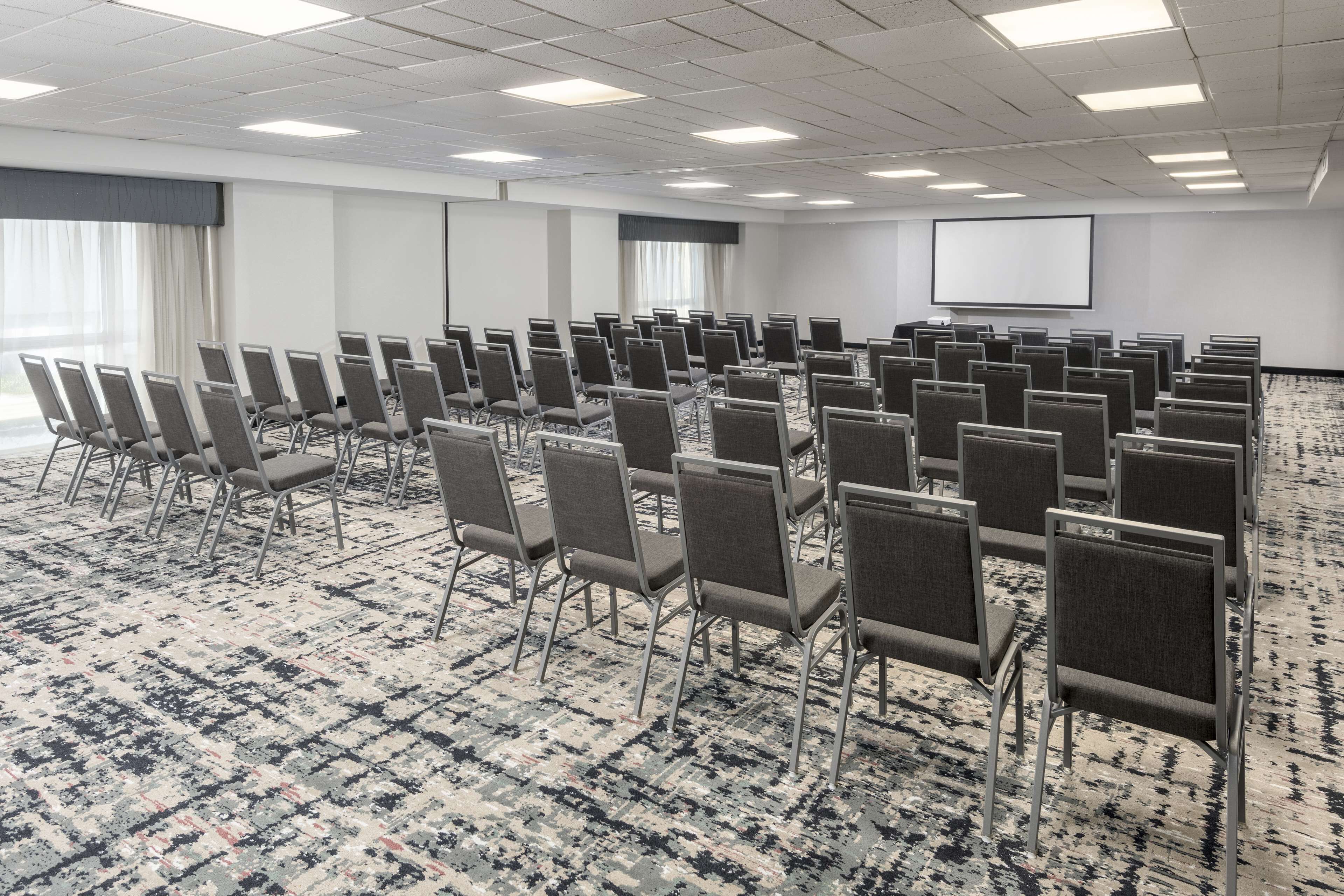 A conference room with rows of chairs, a carpeted floor, a projector screen, and a large window with curtains.
