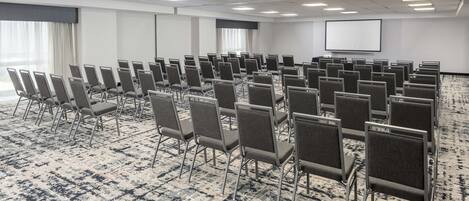 Un salón de conferencias con hileras de sillas, piso alfombrado, pantalla de proyector y un amplio ventanal con cortinas.