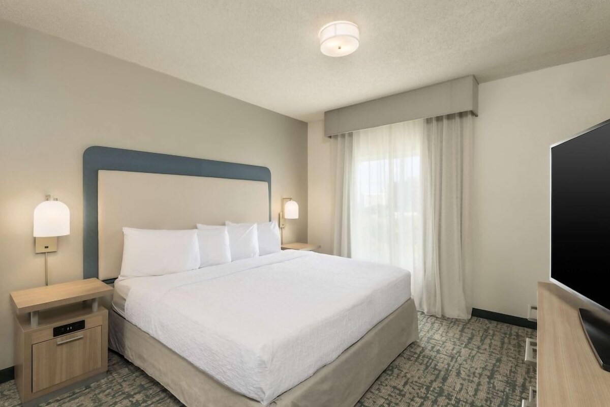 A hotel room with a large bed, two bedside tables, a flat-screen TV, and a window with curtains.