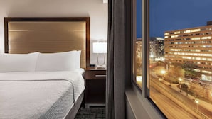 A hotel room with a large bed, a nightstand with a lamp, and a view of a cityscape at night. - Homewood Suites by Hilton Washington, D.C. Downtown (Washington)