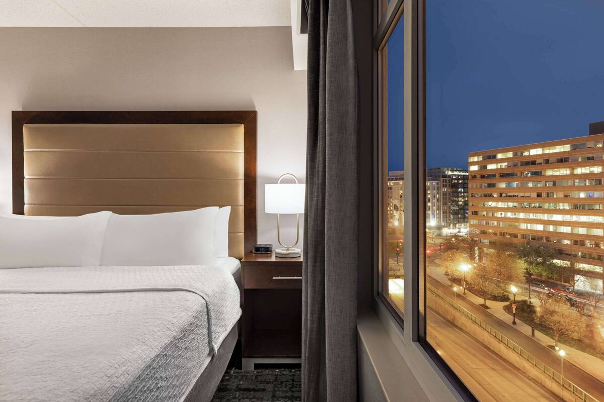 A hotel room with a large bed, a nightstand with a lamp, and a view of a cityscape at night.