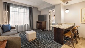 A modern hotel room with a sofa, ottoman, flat-screen TV, dark wood cabinet, and a small kitchen area with a marble countertop and chairs. - Homewood Suites by Hilton Washington, D.C. Downtown (Washington)