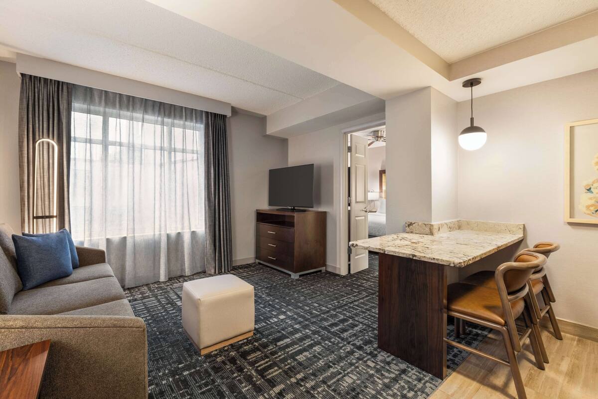 A modern hotel room with a sofa, ottoman, flat-screen TV, dark wood cabinet, and a small kitchen area with a marble countertop and chairs.