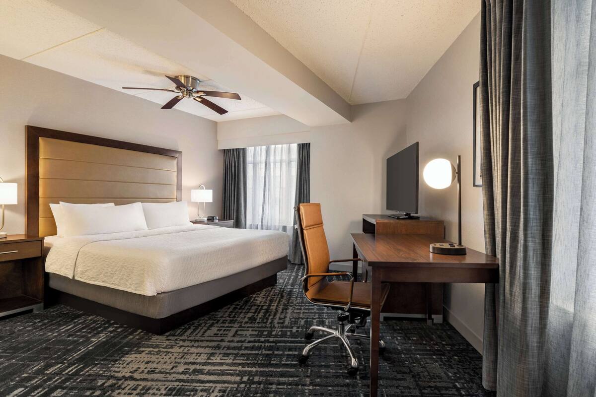 A hotel room with a large bed, a desk, a chair, a television, and a ceiling fan.