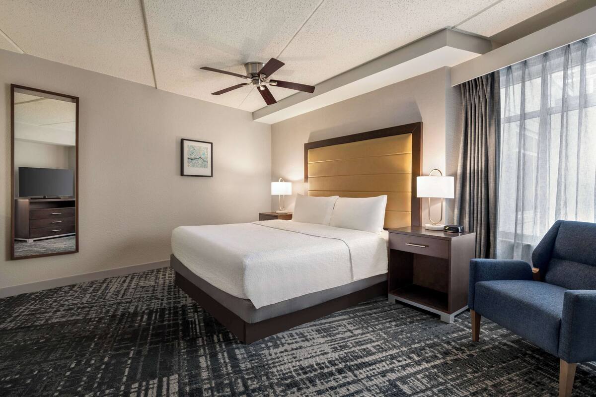 A hotel room with a large bed, a desk with a lamp, a chair, a ceiling fan, and a large window with curtains.