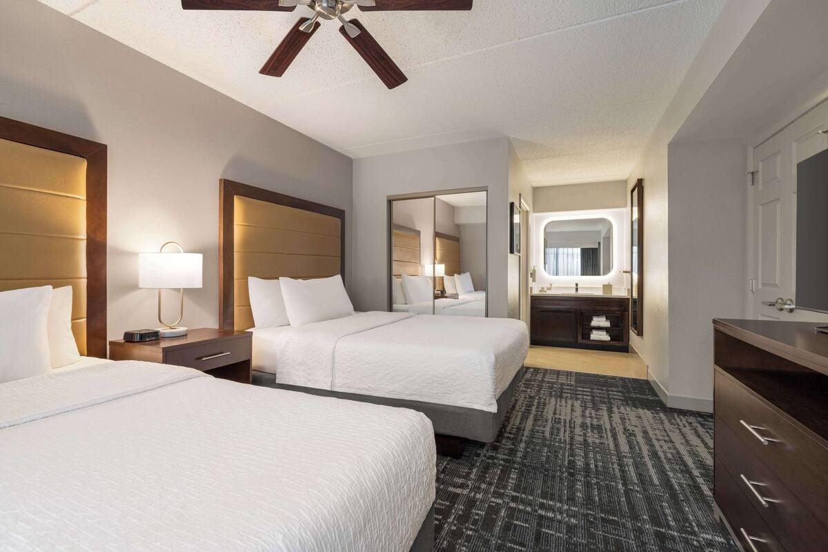 A hotel room with two beds, a ceiling fan, a bedside table with a lamp, a mirror, a dresser, and a bathroom visible through an open door.