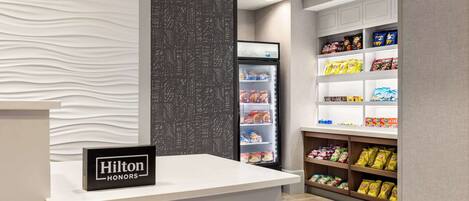 A hotel reception area with a modern design, including a white reception desk, a minibar, and shelves stocked with snacks.