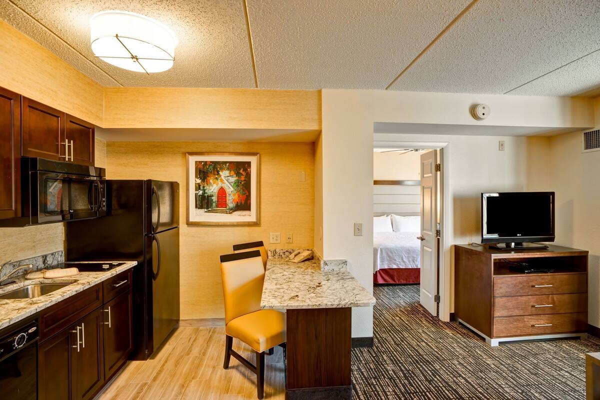 A compact hotel room with a kitchenette, a dining area, a TV, and a bed.
