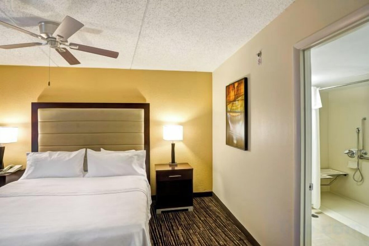 A hotel room with a bed, bedside tables, a ceiling fan, a shower, and a door leading to another room.