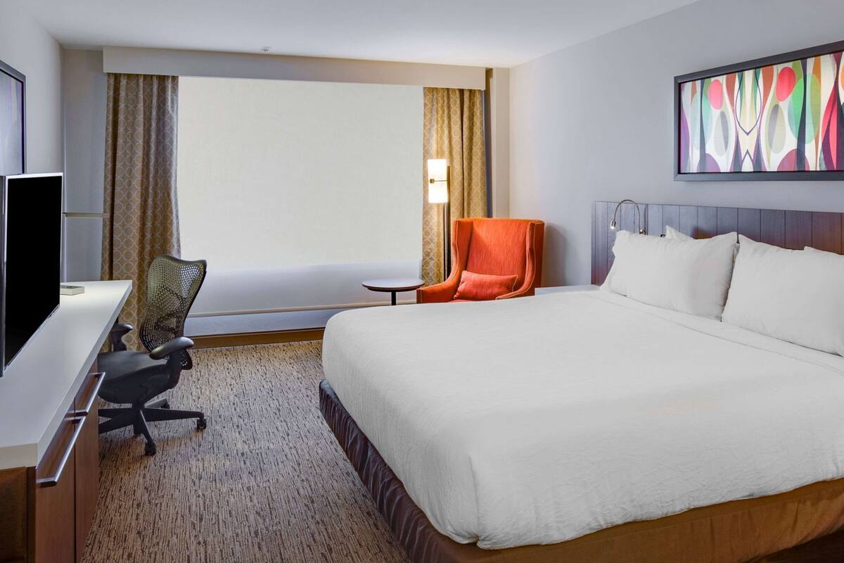 A hotel room with a bed, desk, chair, TV, and a painting on the wall.