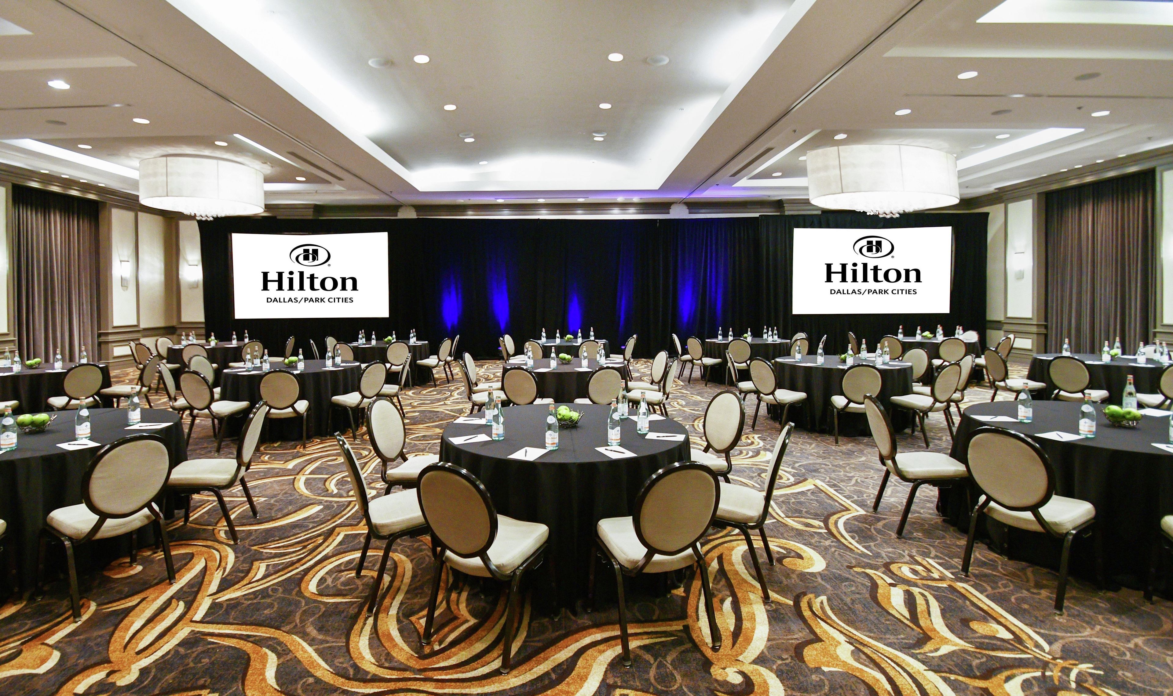 A conference room with multiple tables and chairs, a large screen displaying the Hilton logo, and a carpeted floor.