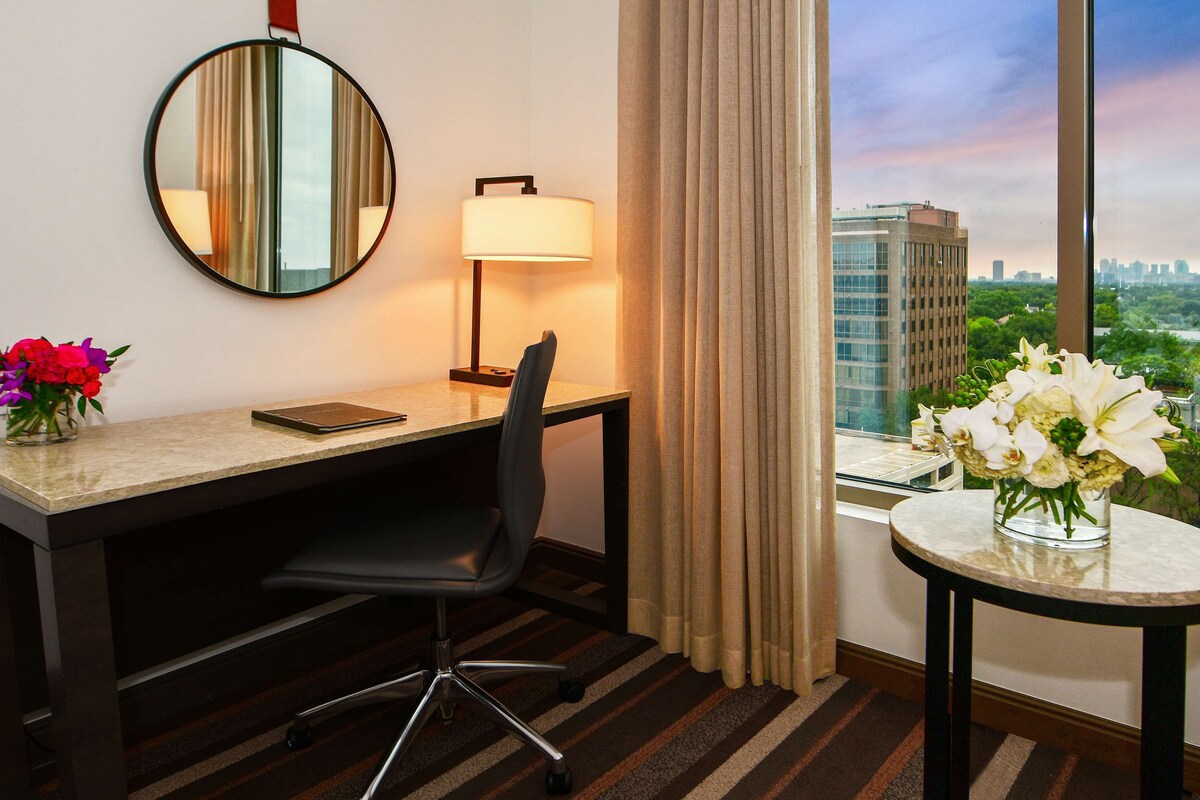 Habitación de hotel con escritorio, silla, lámpara, espejo y vistas de la ciudad a través de amplias ventanas.