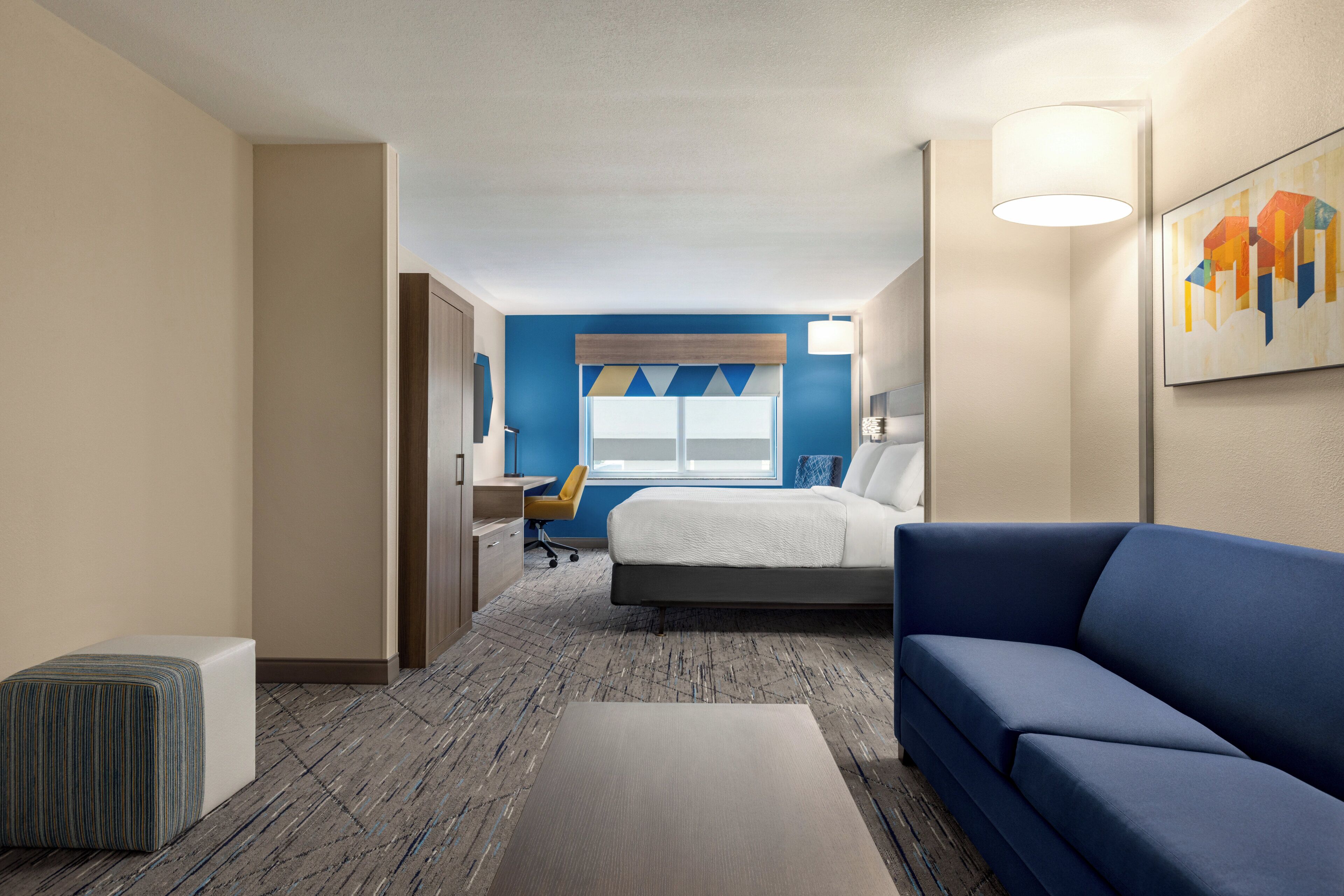 Habitación de hotel con cama, sofá azul, escritorio con silla, ventana con persianas y un cuadro en la pared.