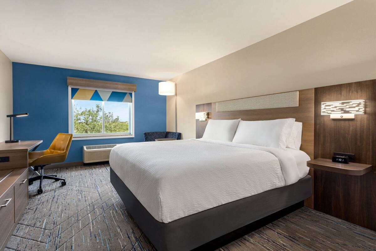 A hotel room with a large bed, a desk, a chair, and a window with a view of greenery.