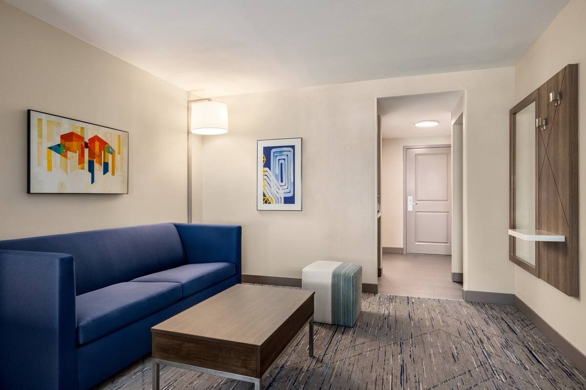A hotel lobby with a blue sofa, a wooden coffee table, a small ottoman, and two framed artworks on the wall.