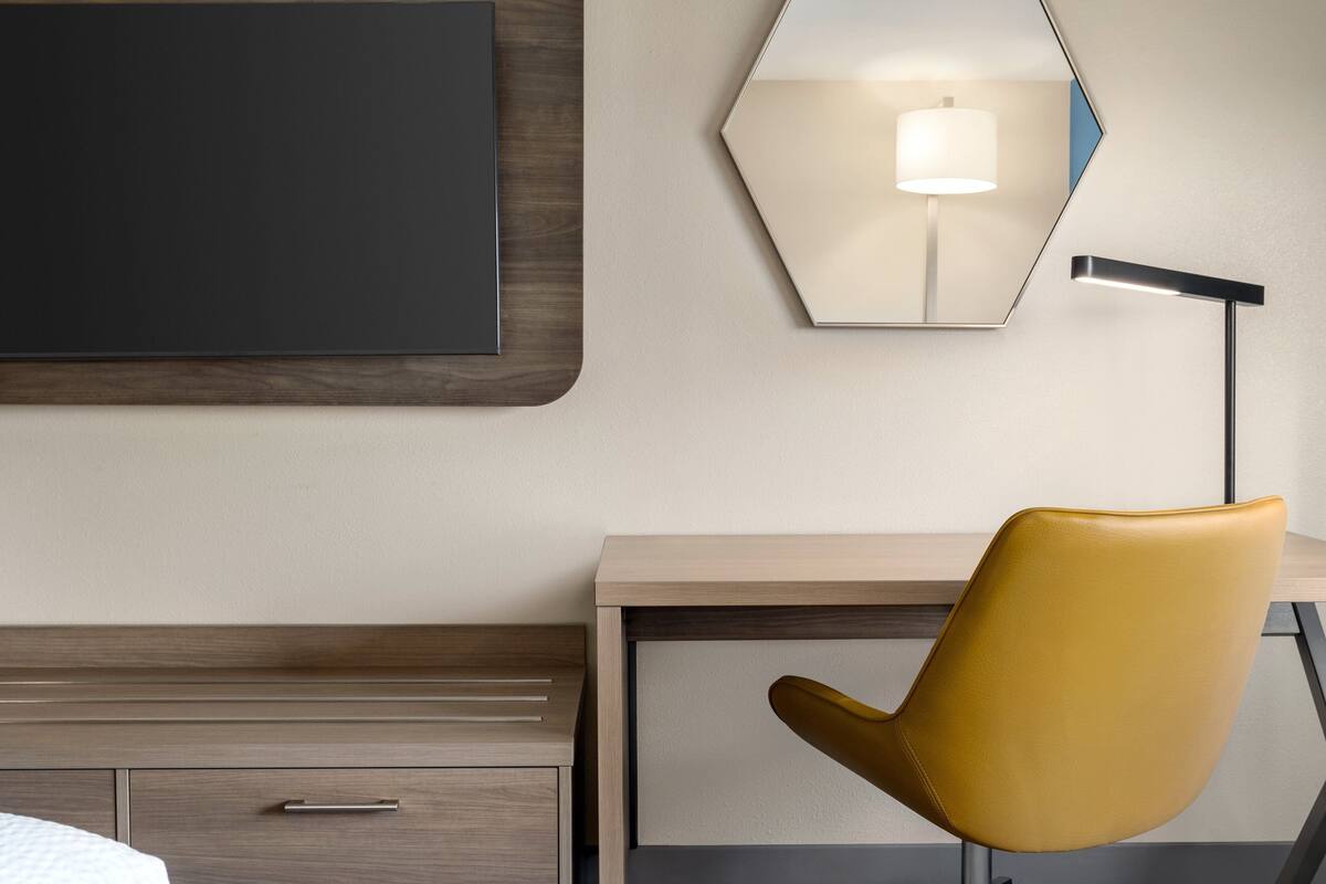 A hotel room with a bed, a desk, a chair, a flat-screen TV, and a wall-mounted lamp.