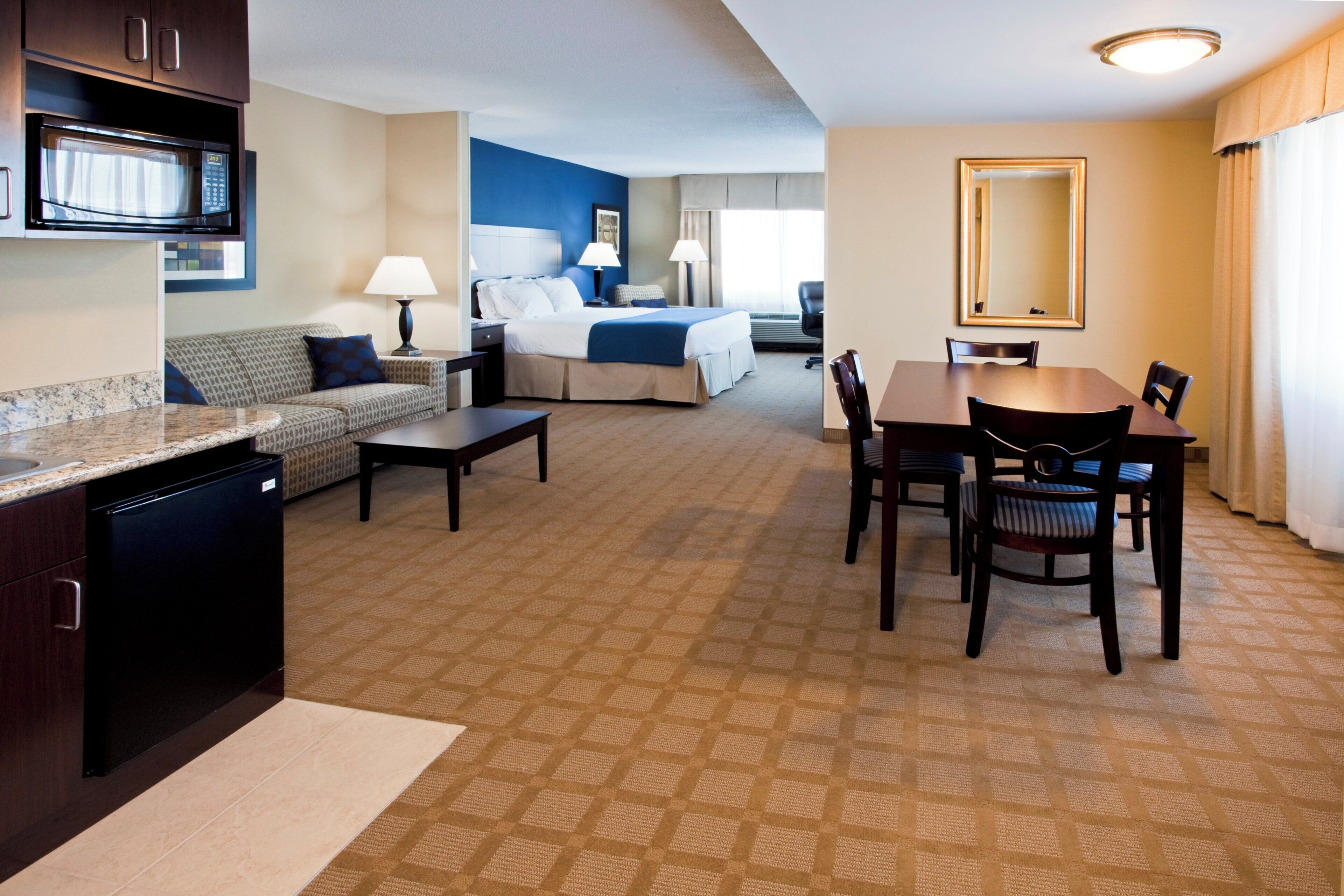 A hotel room with a kitchenette, a bed, a seating area with a sofa and a table, and a dining table with chairs.