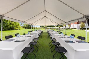 A tented outdoor event space with rows of tables and chairs, a grassy lawn, and a building in the background.