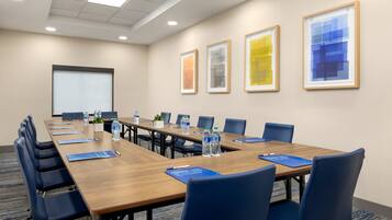 Une salle de conférence avec une longue table, des chaises bleues, des bouteilles d’eau et des œuvres d’art encadrées accrochées au mur.