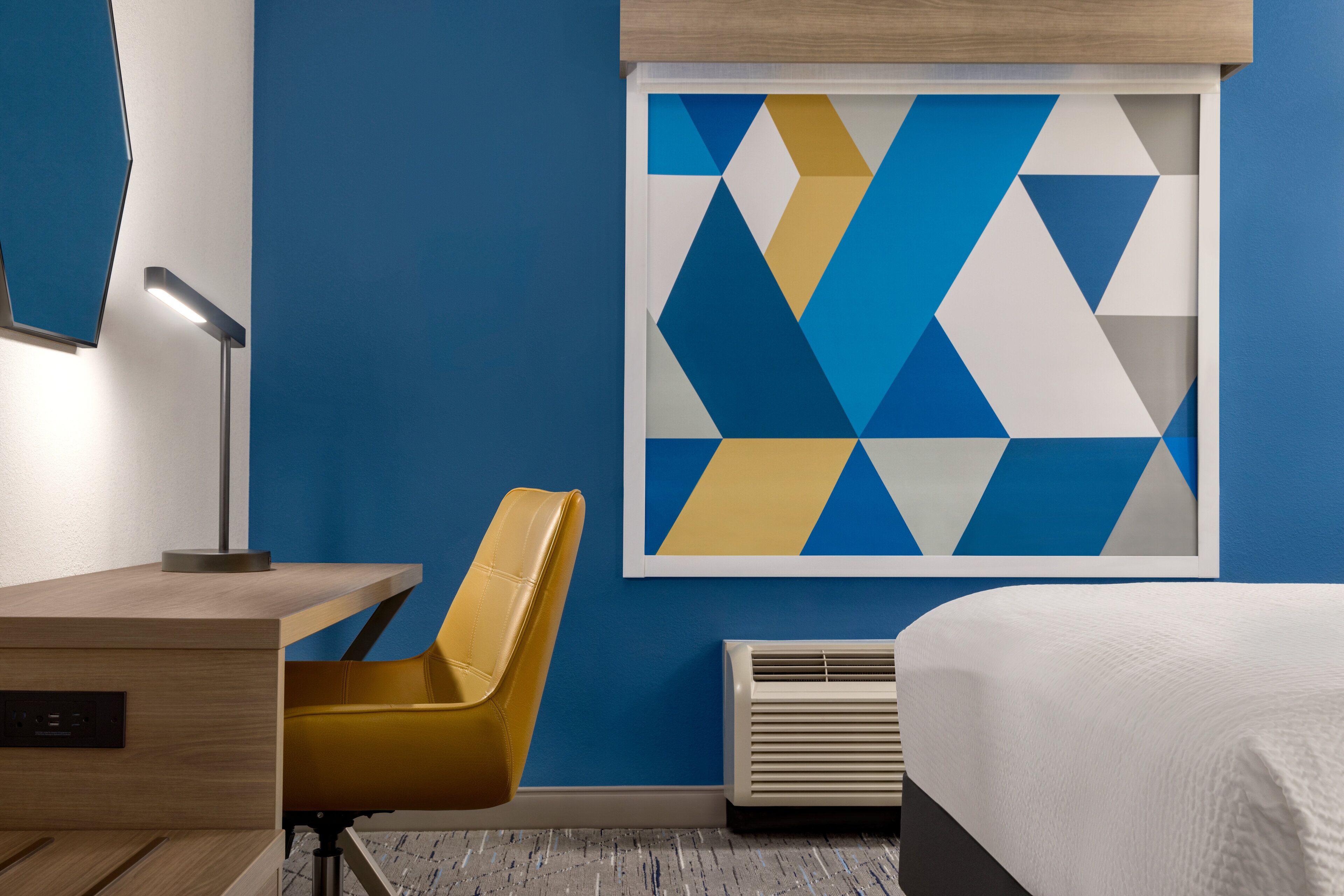 Habitación de hotel con cama, escritorio con lámpara, silla, aire acondicionado instalado en la pared y una obra de arte con motivos geométricos.