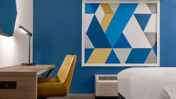 Habitación de hotel con cama, escritorio con lámpara, silla, aire acondicionado instalado en la pared y una obra de arte con motivos geométricos.