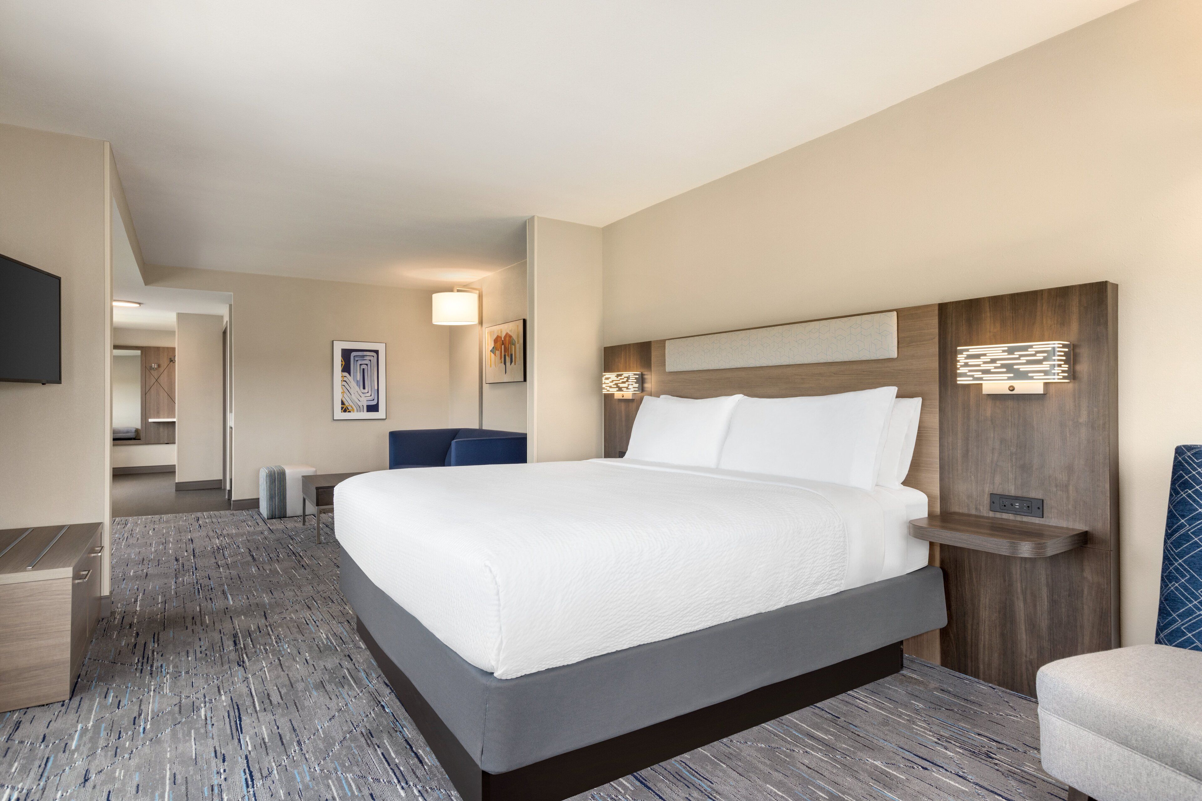 A hotel room with a large bed, a desk, a chair, a TV, and a wall-mounted shelf.