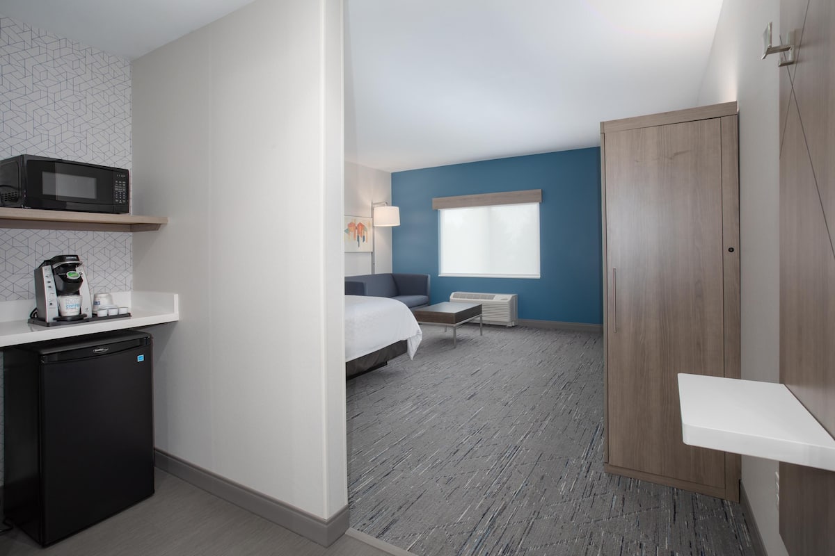 A compact hotel room with a kitchenette, a bed, a bedside table, a window, and a wardrobe.
