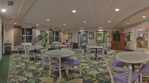 A hotel dining area with multiple round tables and chairs, a patterned carpet, and a ceiling with recessed lighting.