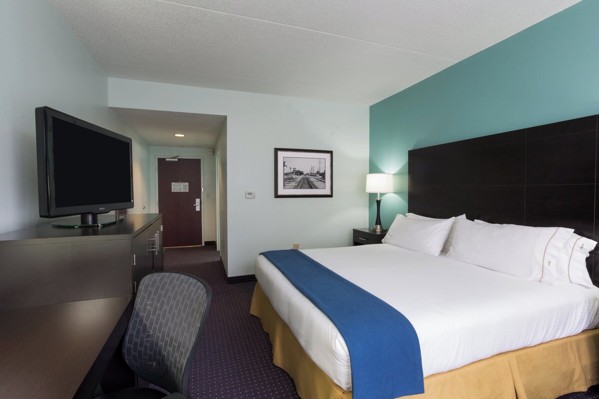 A hotel room with a bed, a desk with a television, a chair, a picture on the wall, and a nightstand with a lamp.