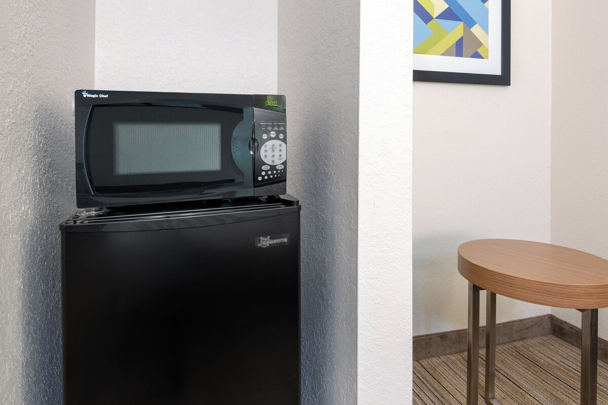 Ein schwarzer Kühlschrank mit einer Mikrowelle obenauf, ein gerahmtes Bild an der Wand und ein Holzschemel.