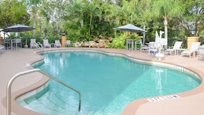 A swimming pool with a 3 FT depth marker, surrounded by lounge chairs and umbrellas, with a rocky wall and lush greenery in the background.