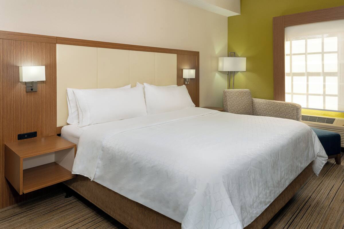 Ein Hotelzimmer mit einem großen Bett, zwei Nachttischen, einem Sessel und einem Fenster mit Jalousien.