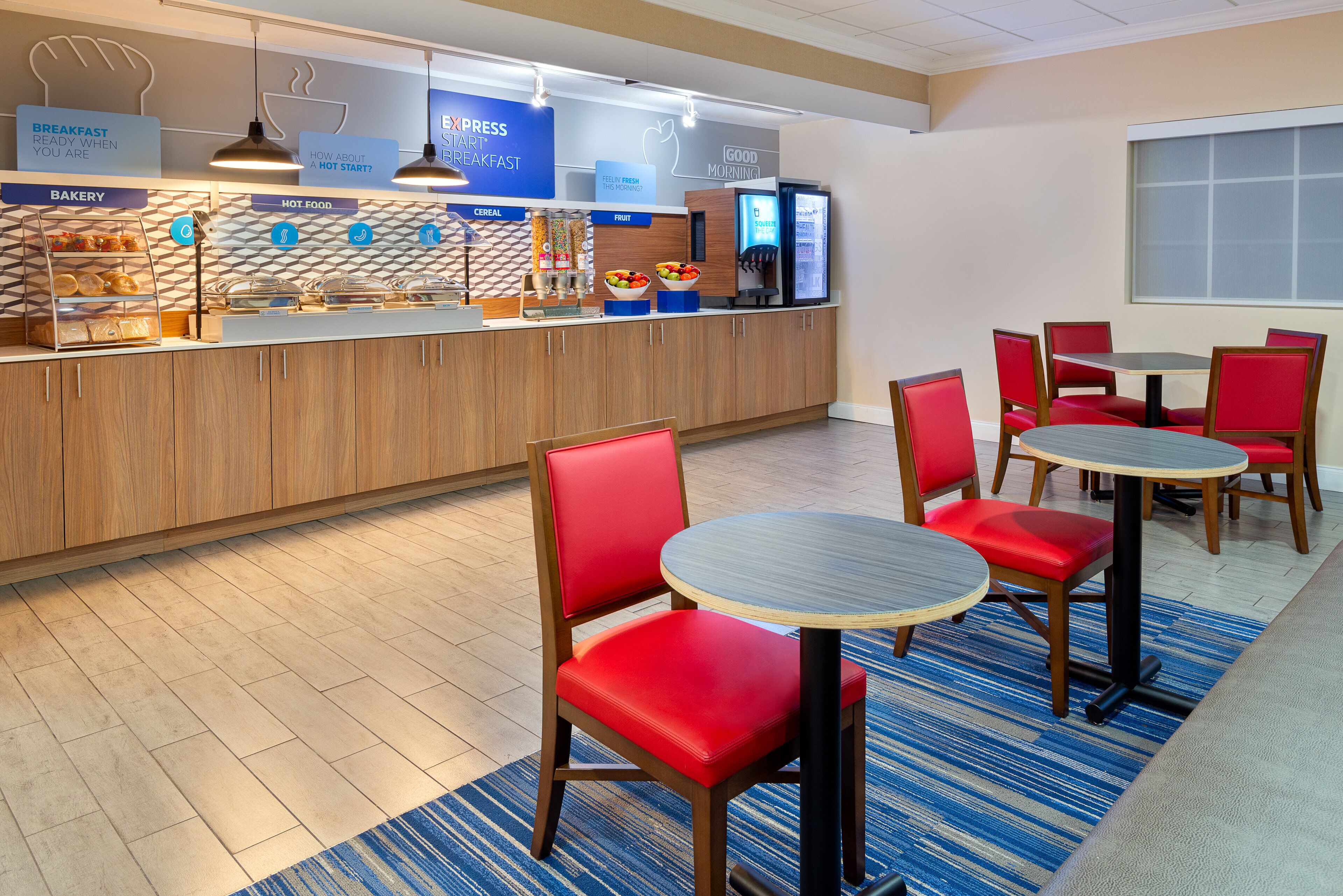 A breakfast bar with bakery, hot food, cereal, and fruit options, and a seating area with red chairs and round tables.