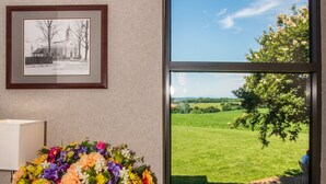 Una habitación con un amplio ventanal, un cuadro enmarcado en la pared y una mesa con un jarrón de flores.