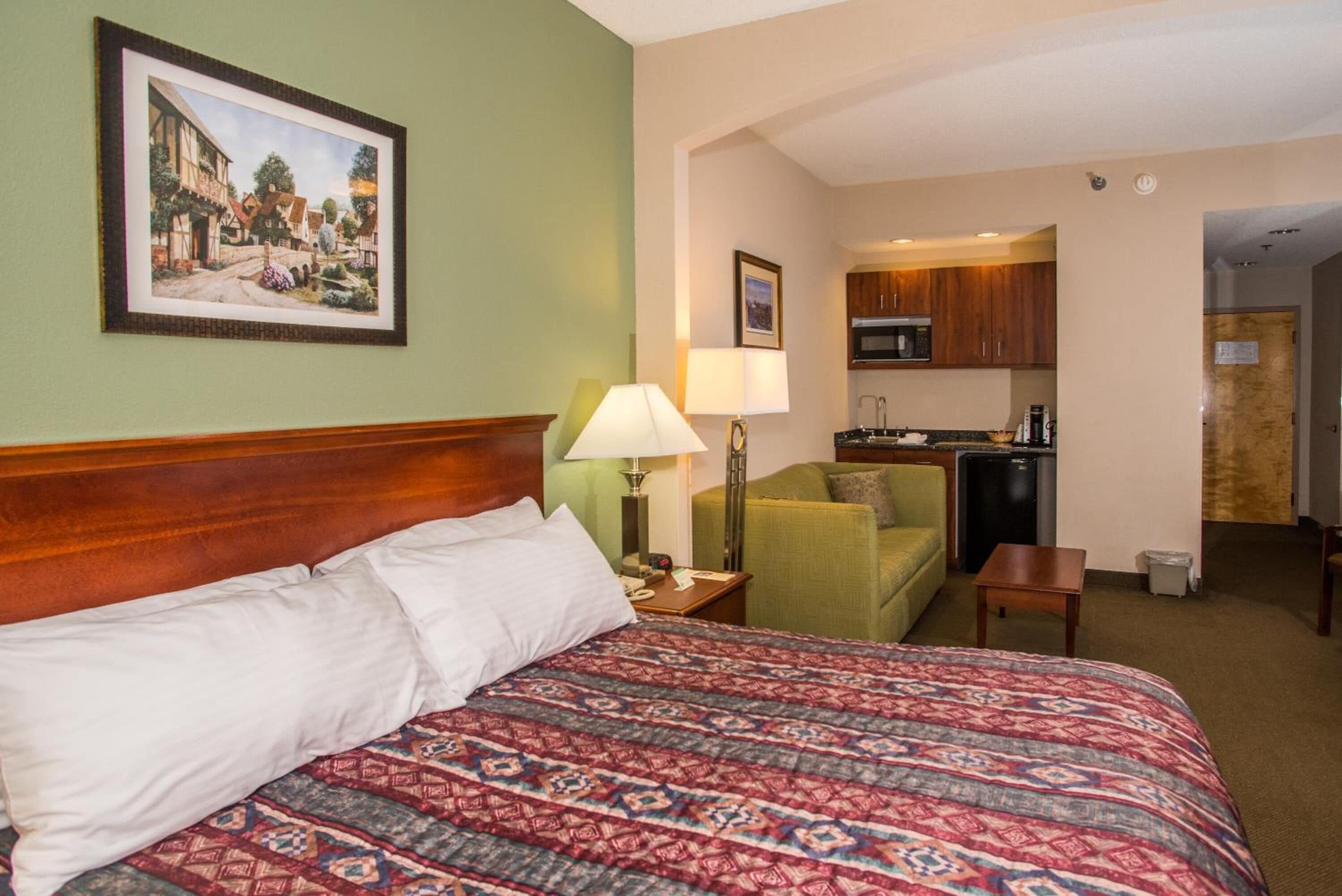 A hotel room with a large bed, a green armchair, a small table, a lamp, a kitchenette, and a framed picture on the wall.