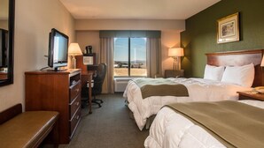 Habitación de hotel con dos camas, un escritorio con computadora, una silla, televisor y un amplio ventanal con vistas al exterior.