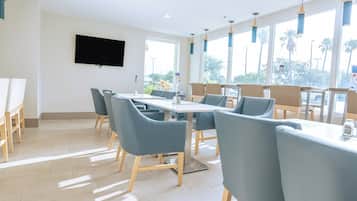 Uma área de jantar moderna com uma mesa comprida e várias cadeiras, janelas amplas com vista para palmeiras e uma TV de tela plana na parede.
