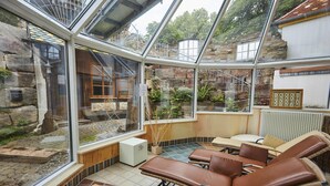 Een wintertuin met glazen wanden, stenen muren en een open haard van natuursteen. Er staan comfortabele fauteuils en een klein tafeltje.