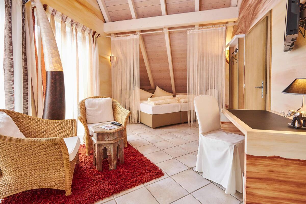 A cozy room with a high ceiling, a bed, two wicker chairs, a small wooden table, and a desk with a lamp.