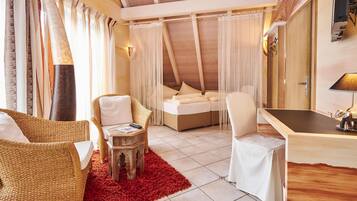 A cozy room with a high ceiling, a bed, two wicker chairs, a small wooden table, and a desk with a lamp.