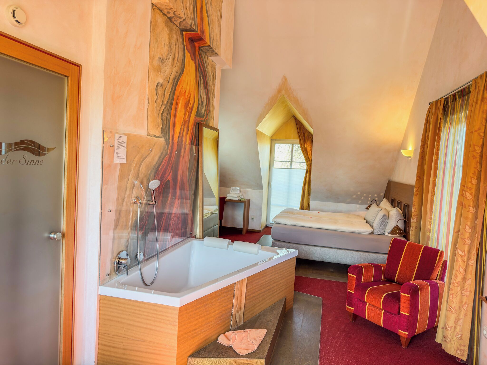 A hotel room with a large bathtub, a bed, a red armchair, and a wooden vanity.