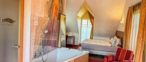A hotel room with a large bathtub, a bed, a red armchair, and a wooden vanity.
