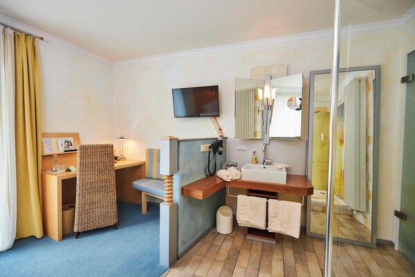 A hotel room with a wooden desk, a chair, a television, a mirror, a sink, and a shower area.